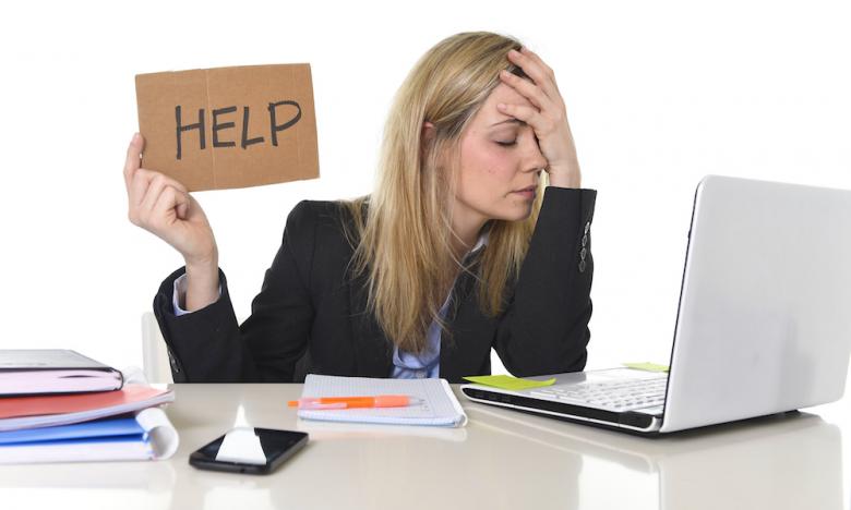 Best online business ideas for beginners - Lady working at her desk on her laptop computer holding her head in one hand and a cardboard sign that says "help" in the other.