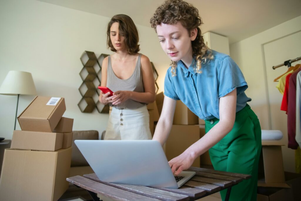 Online Business Ideas for Beginners - two ladies, one working at the desk on her laptop computer and the other packing boxes for shipping